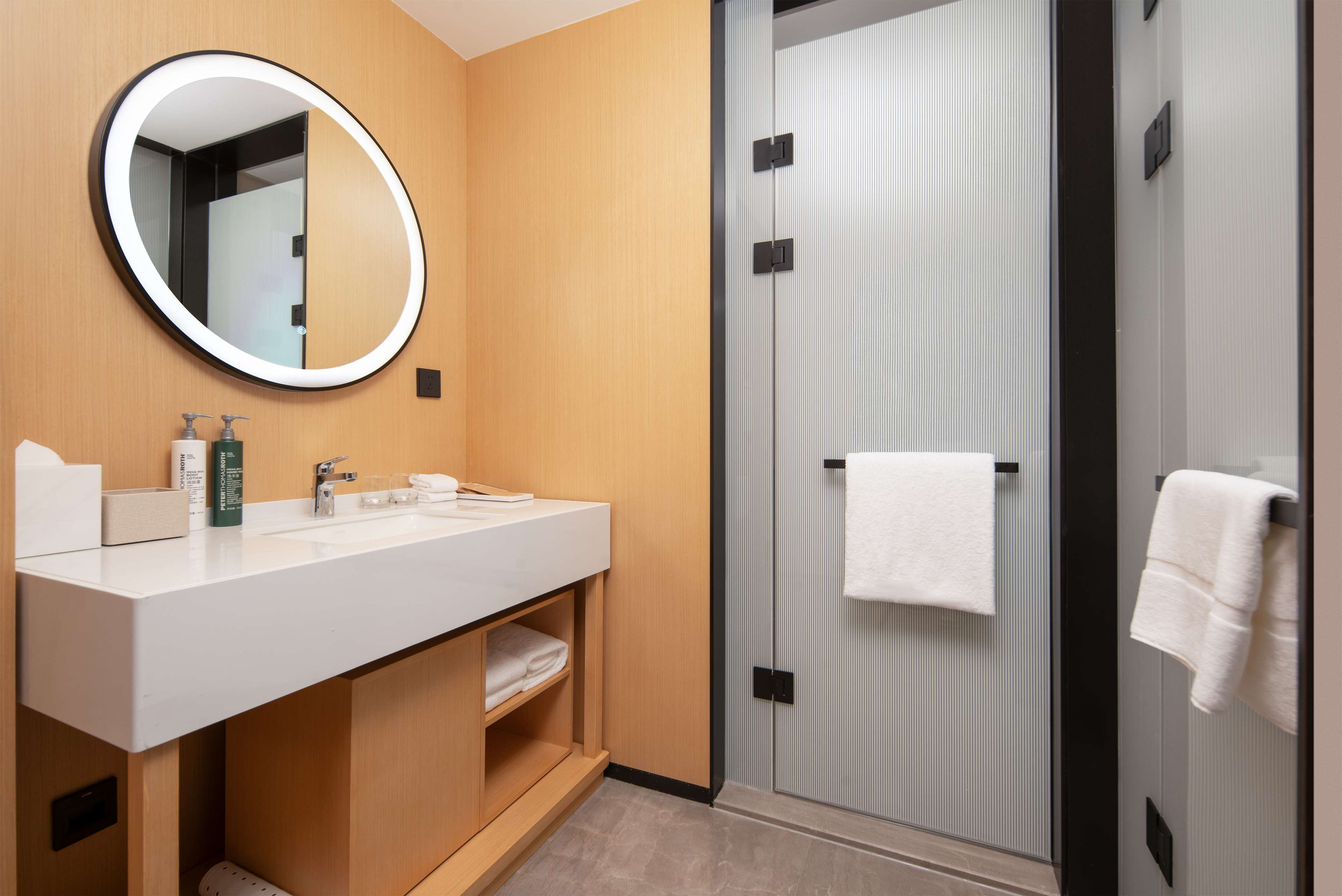 Un baño moderno con espejo redondo, lavamanos blanco y ducha con mampara de vidrio.