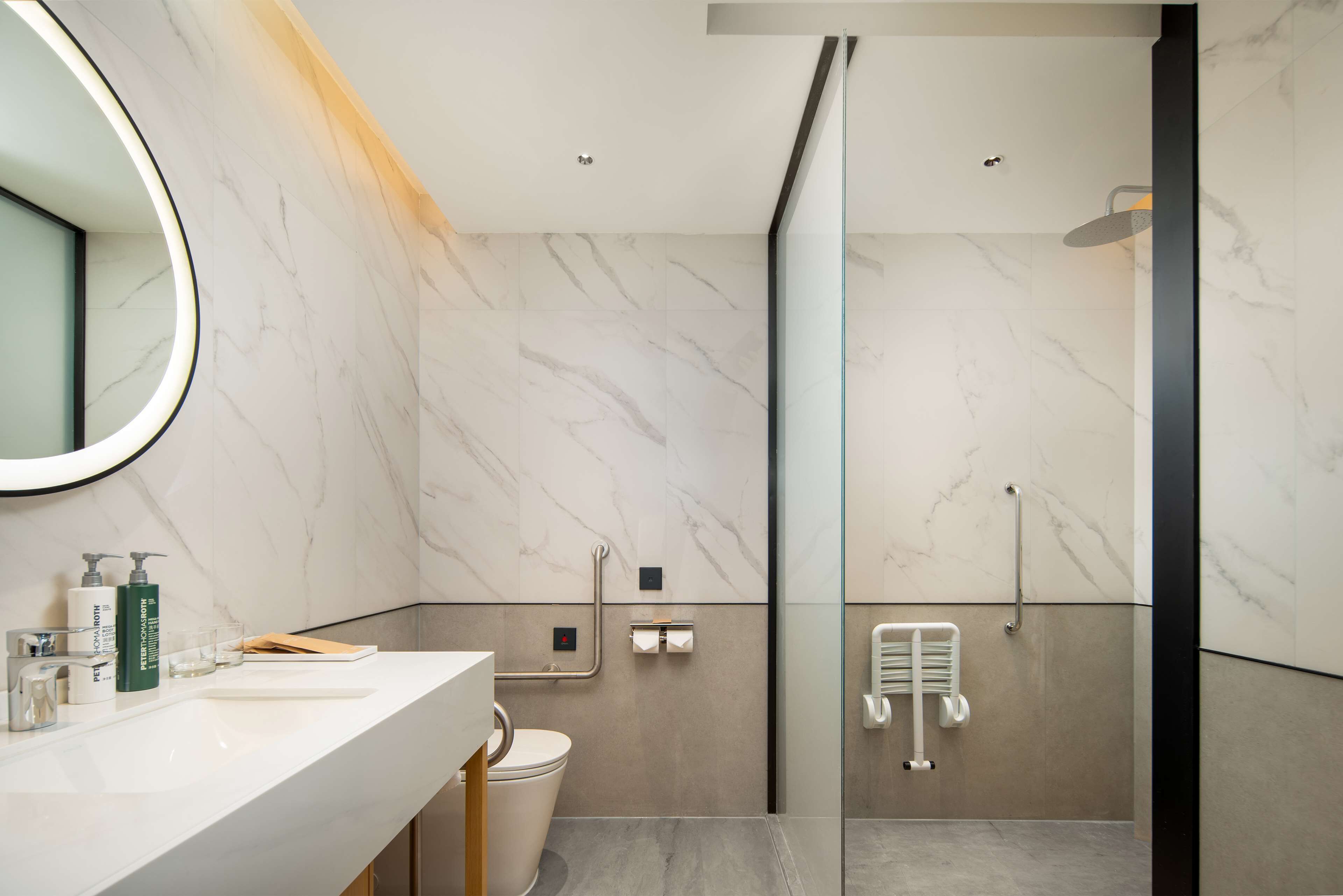 Un baño moderno con pared de mármol, cabina de ducha con mampara de vidrio y lavamanos blanco con artículos de aseo.