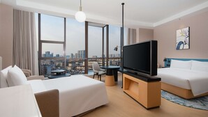Une chambre d’hôtel avec un grand lit, un lit plus petit, une télévision, un bureau et une chaise. Il y a une grande fenêtre donnant sur la ville.