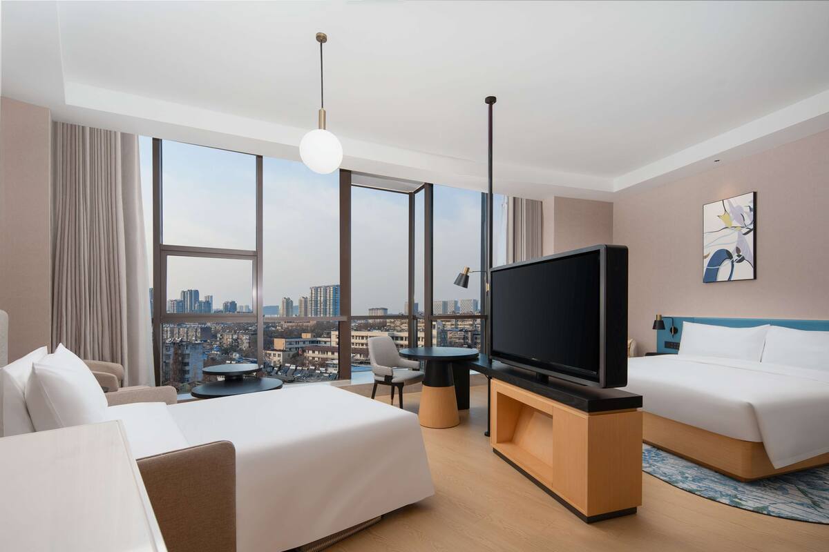 Une chambre d’hôtel avec un grand lit, un lit plus petit, une télévision, un bureau et une chaise. Il y a une grande fenêtre donnant sur la ville.