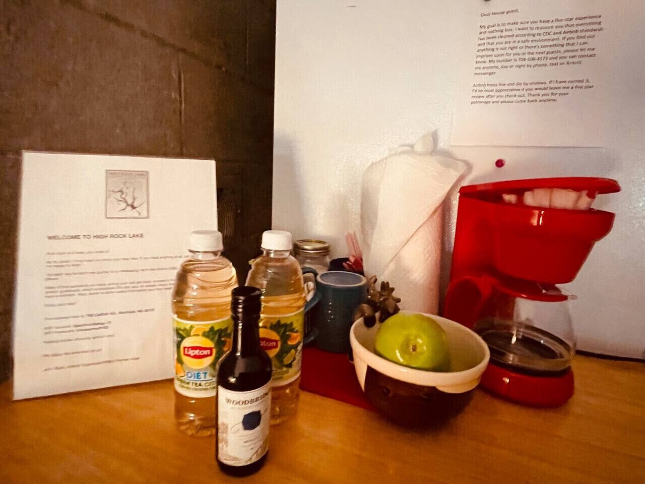 Una mesa con un menú, una botella de té Lipton, una botella de agua tónica sin azúcar, una botella de agua, un bol con una manzana y una cafetera roja.