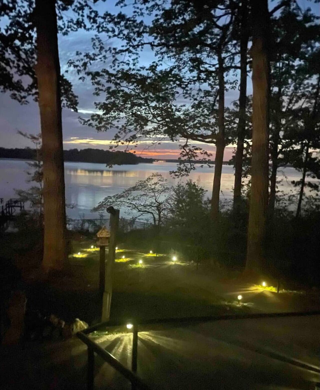 Maisema järven rannalta hämärän aikaan, jossa näkyy puita ja laituri.