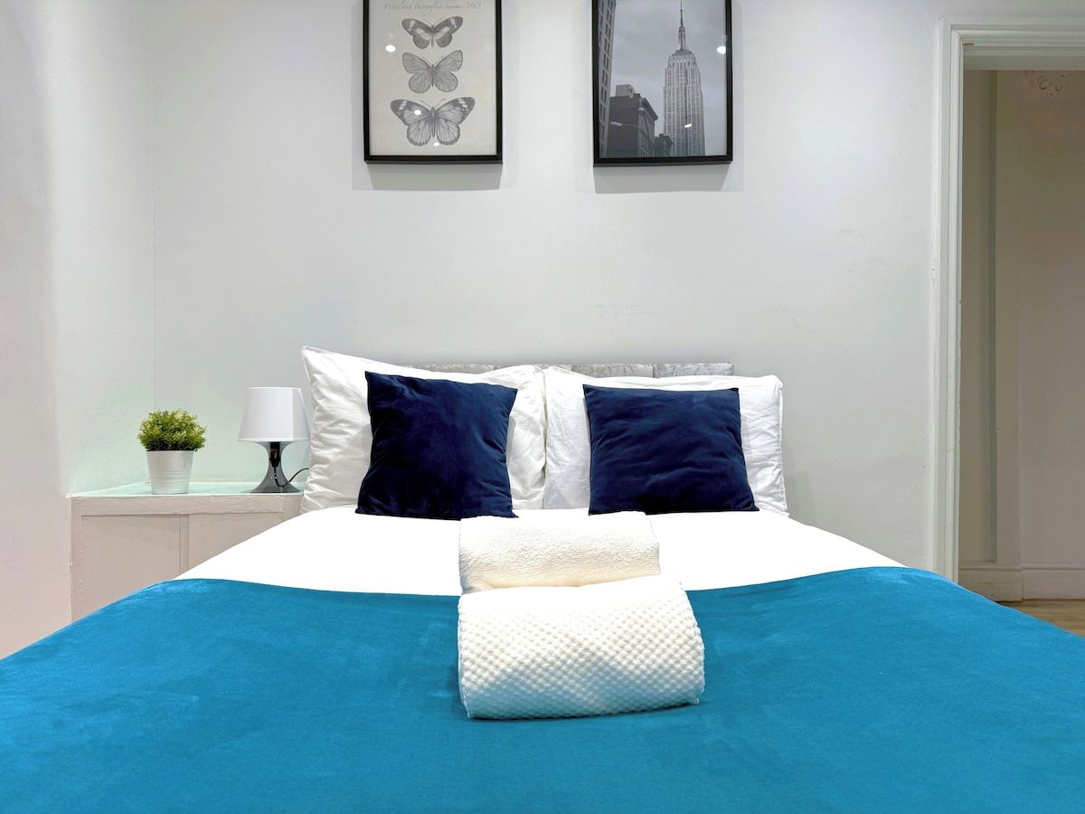 A bed with a blue bedspread, white pillows, and a white pillow on the side. There is a framed picture of butterflies on the wall above the bed.