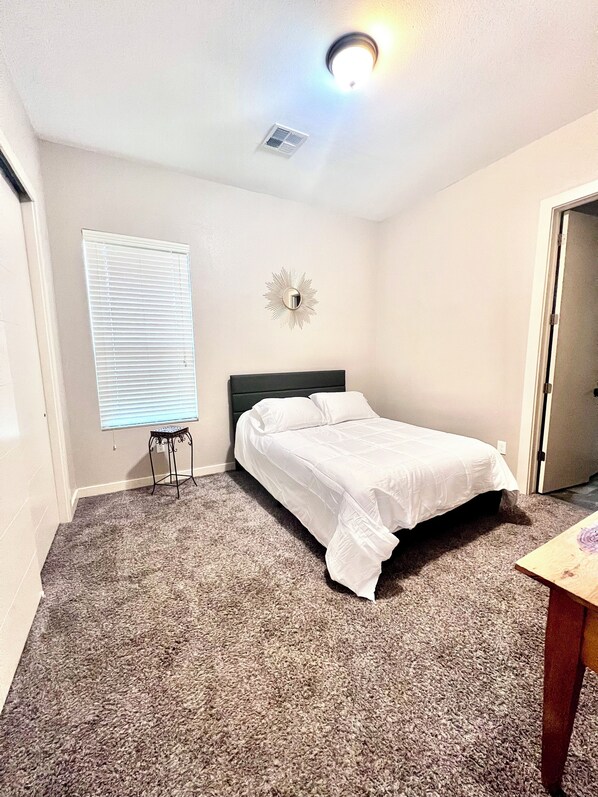 A bedroom with a single bed, a small table, a wall-mounted light fixture, a window with blinds, and a door. - Modern Mountainside Pool House with Sunset Views on West Side (El Paso)