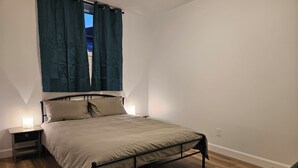 Una cama individual con estructura metálica, dos mesitas de noche y una ventana con cortinas.