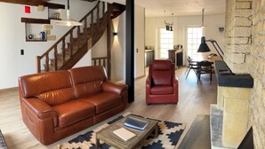 A spacious living room with a leather sofa, a red armchair, a wooden coffee table, and a stone fireplace. A staircase leads to a kitchen area in the background. - Charming house with panoramic terrace in Domme - Sarlat (Domme)