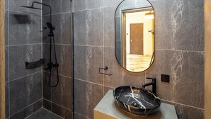 A modern bathroom with a glass-enclosed shower, a marble-tiled wall, a round mirror, and a black marble sink. - Reoky Pads - Redefining comfort and luxury. (Accra)