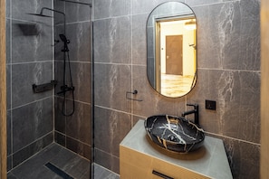 A modern bathroom with a glass-enclosed shower, a marble-tiled wall, a round mirror, and a black marble sink. - Reoky Pads - Redefining comfort and luxury. (Accra)