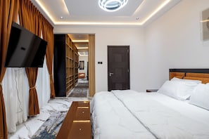 A modern hotel room with a large bed, a flat-screen TV mounted on the wall, and a walk-in closet with shelves. - Reoky Pads - Redefining comfort and luxury. (Accra)