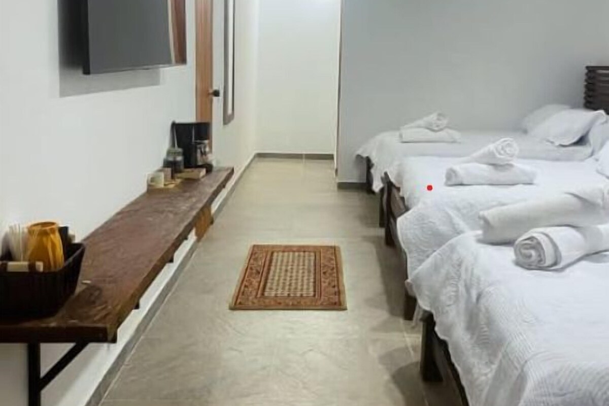 Una habitación de hotel con un largo banco de madera, un televisor de pantalla plana montado en la pared, una cama con ropa blanca y una pequeña mesa con cafetera.