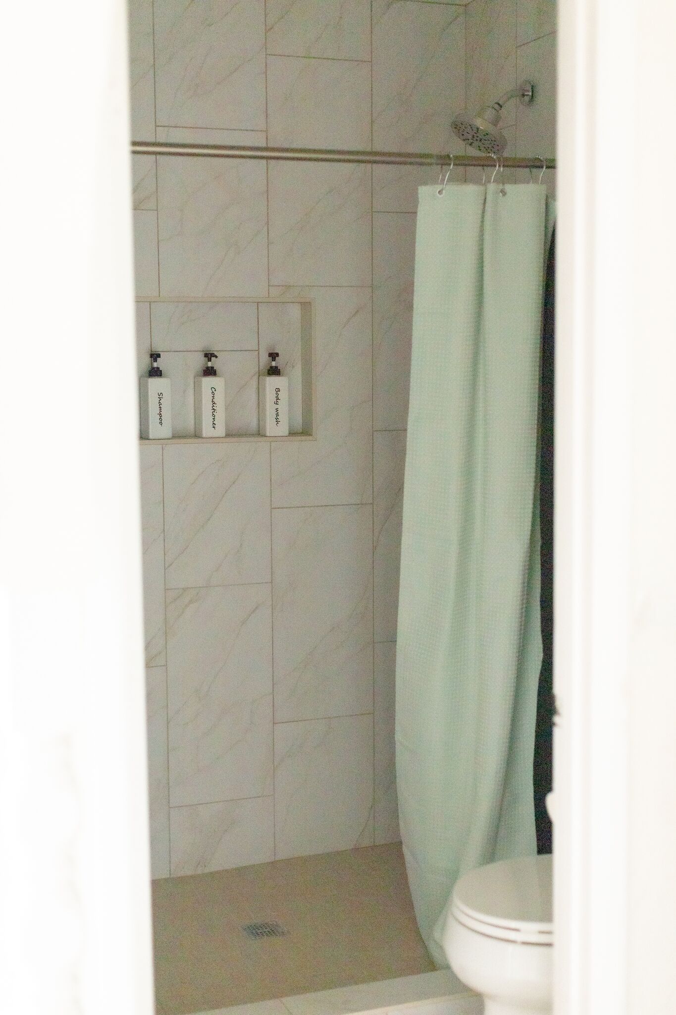 A bathroom with a shower, a toilet, and three bottles of toiletries on a shelf.