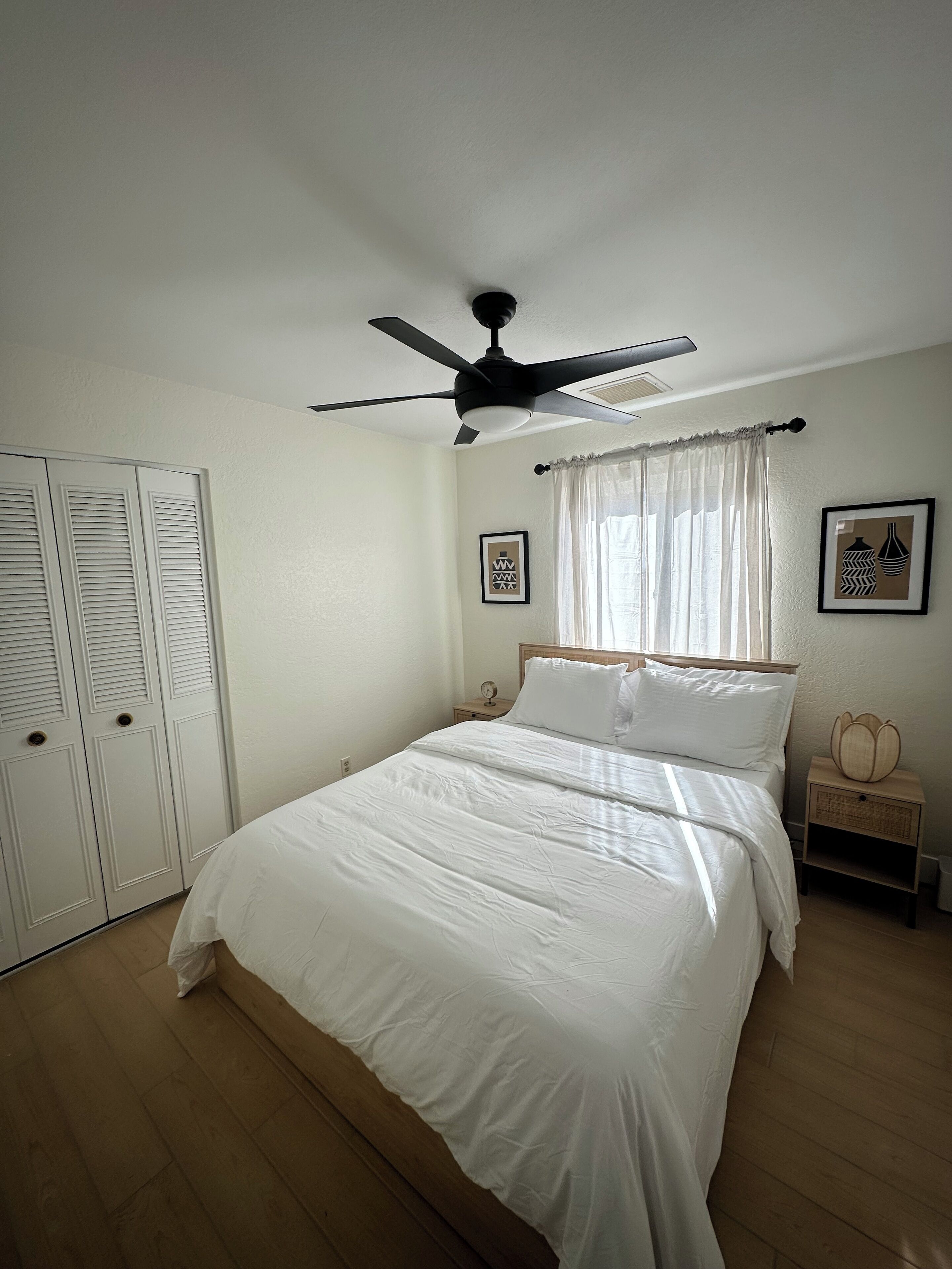 Ein Schlafzimmer mit einem Bett, Deckenventilator, Nachttisch und Vorhängen am Fenster.