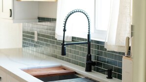 Eine Küche mit weißer Arbeitsplatte, einem schwarzen Wasserhahn und einem Fenster mit transparenten Vorhängen.