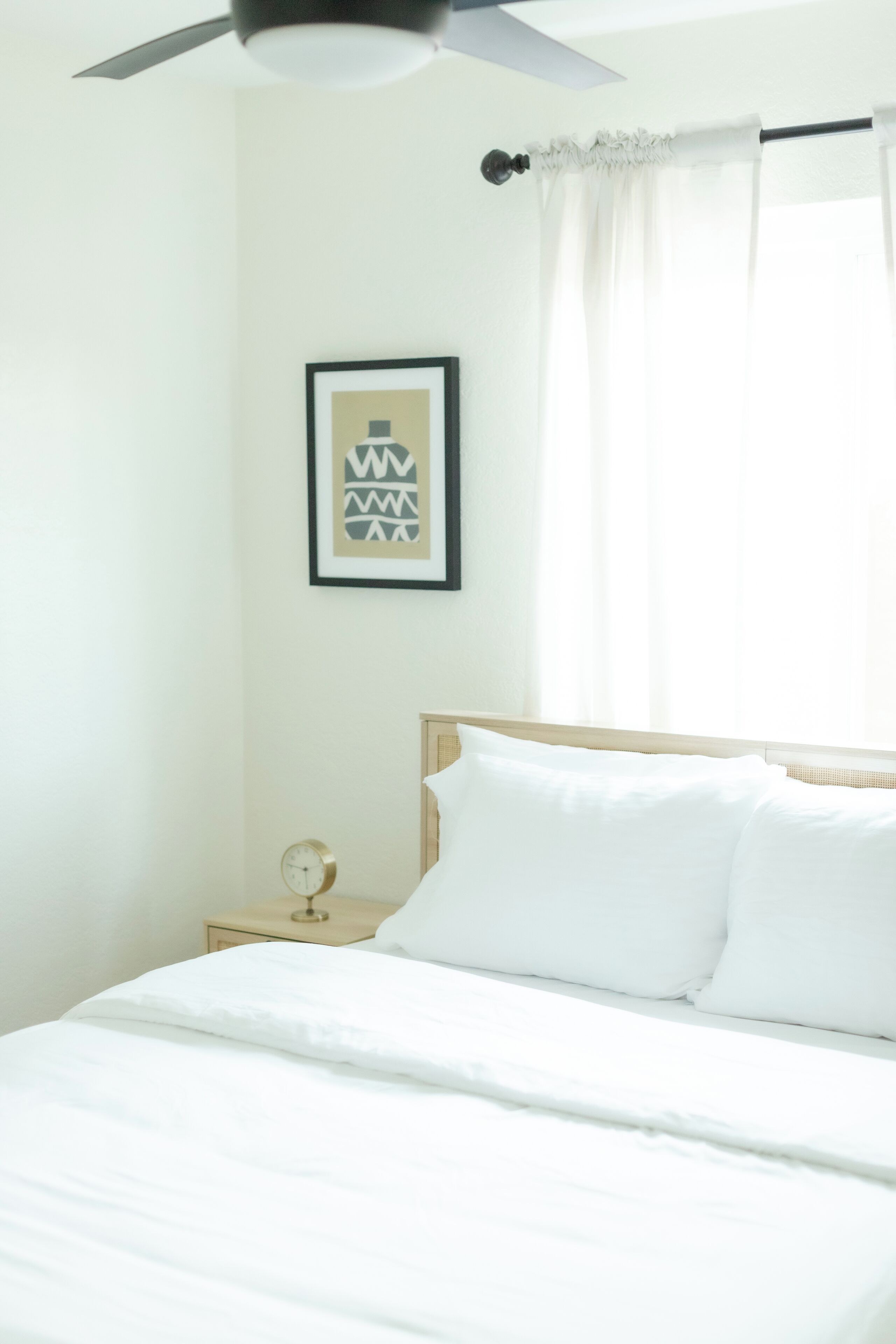 Ein Schlafzimmer mit Bett, Deckenventilator, einem gerahmten Bild an der Wand und einem Nachttisch mit einer Uhr.