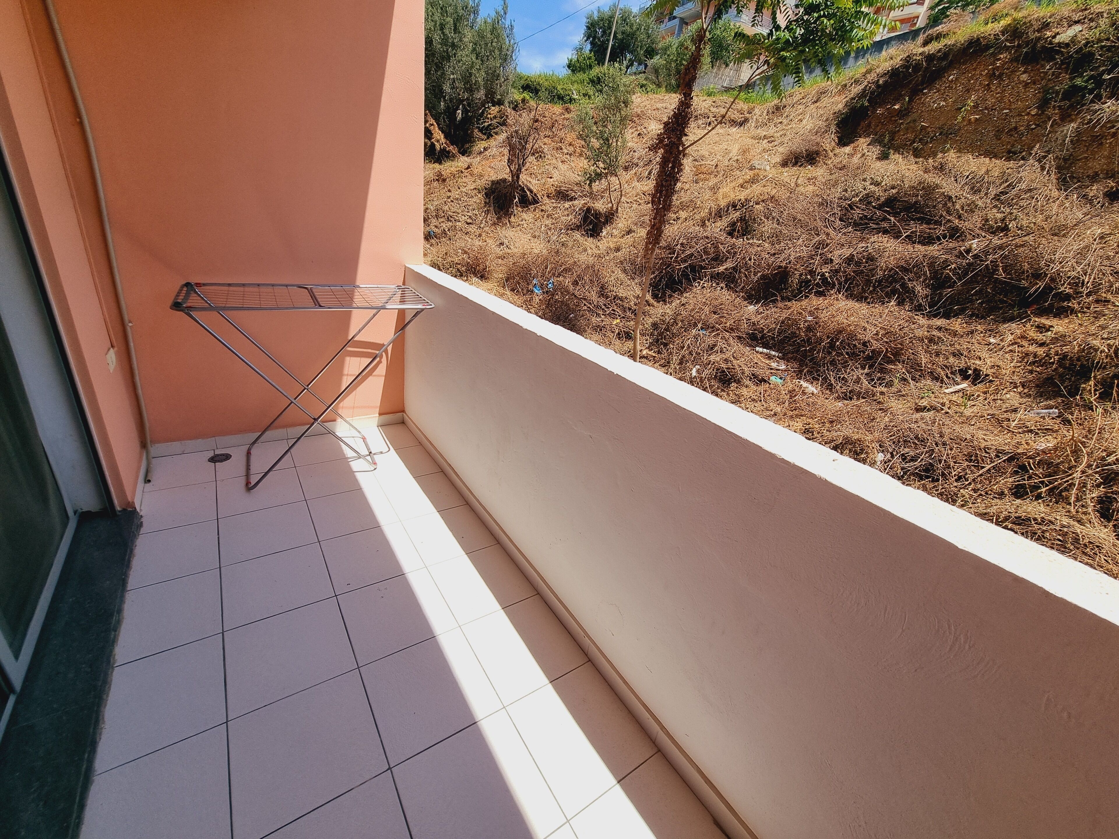 En balkon med et metalrørstativ, flisegulv og udsigt over tørt vegetation og bakkelandskab.