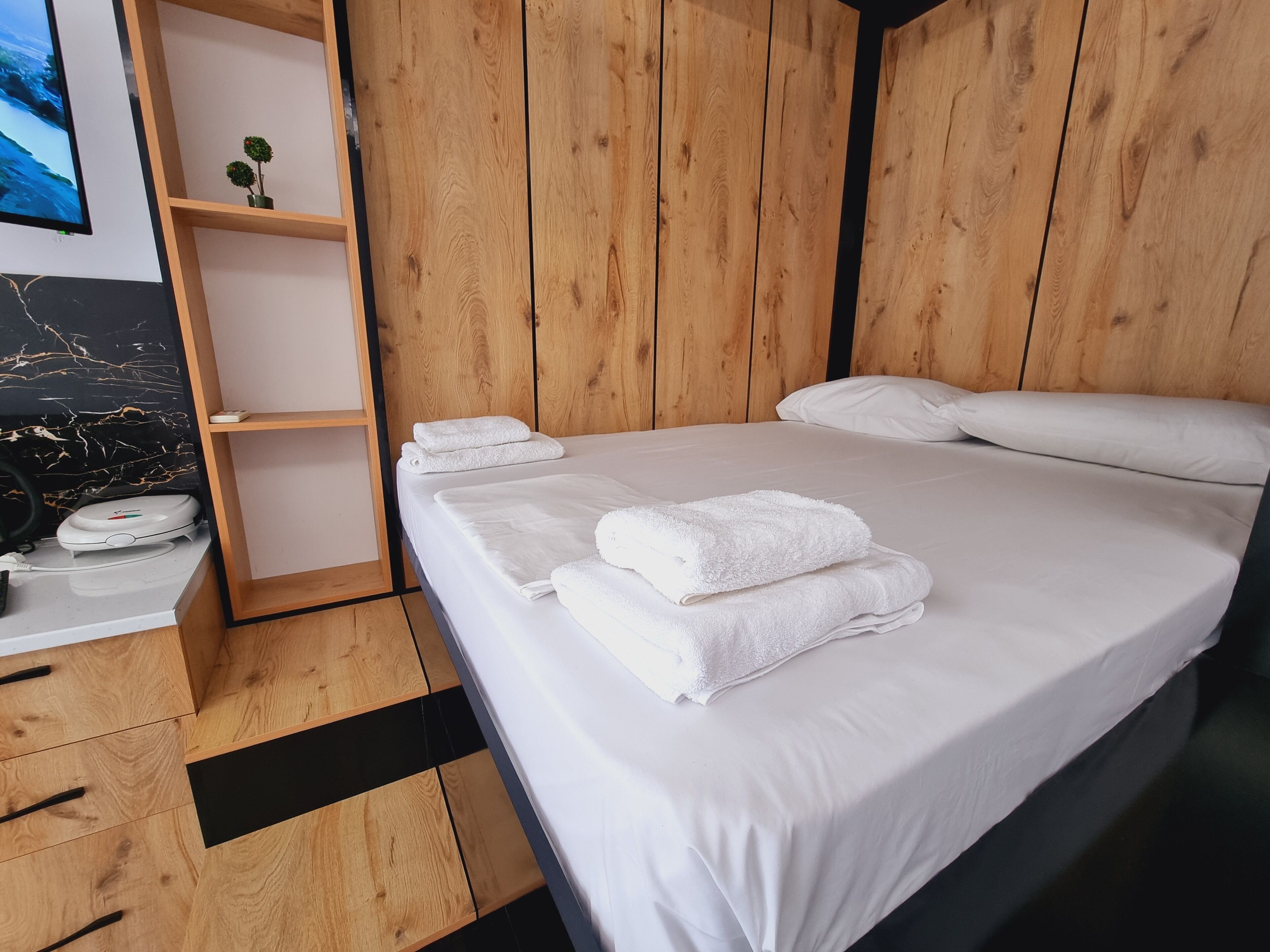 Un lit bien fait, avec du linge de lit et des serviettes blanches, une table de chevet en bois sur laquelle se trouve une balance, une petite étagère avec une plante et une tête de lit en bois.