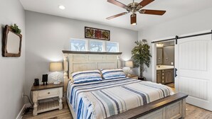 Un dormitorio con cama, mesitas de noche, ventilador de techo, espejo y puerta que da al baño.