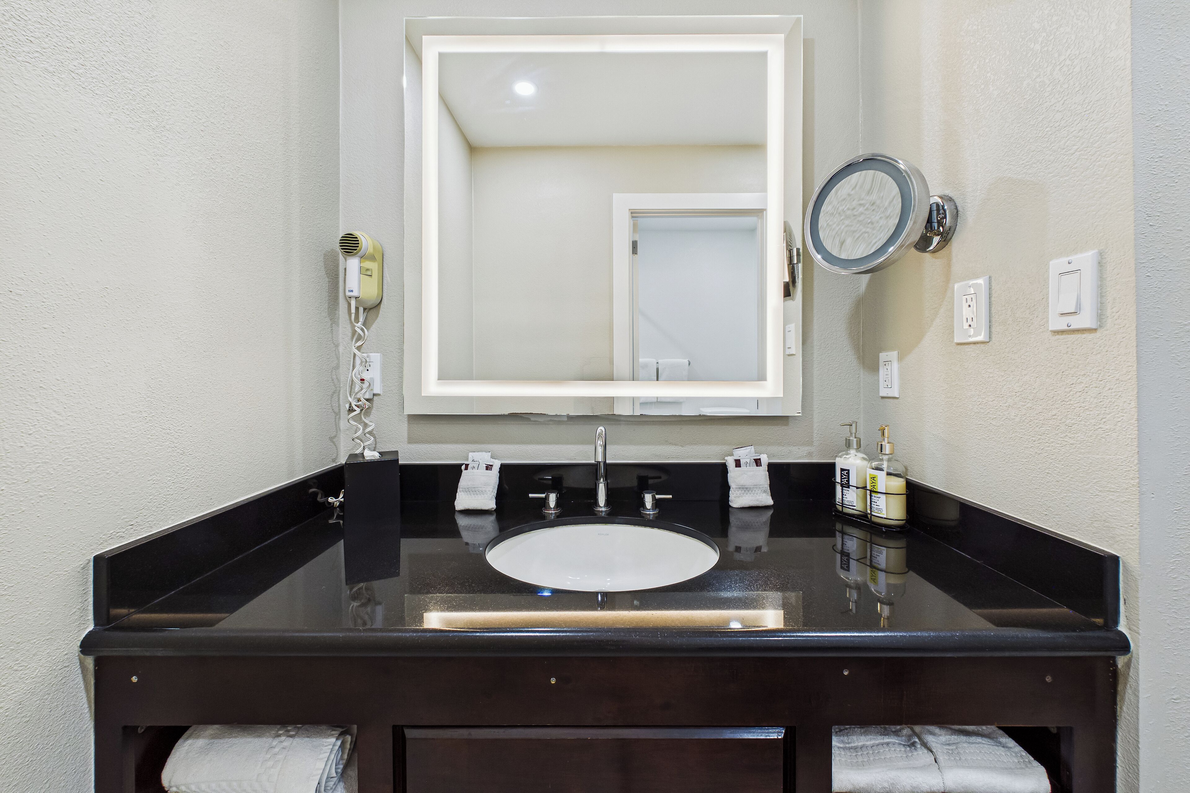 A bathroom with a black sink, a mirror, a hairdryer, and two bottles of shampoo.