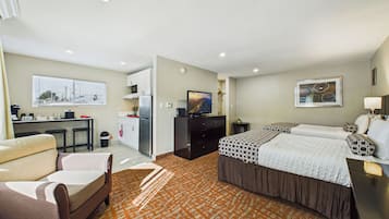 A hotel room with a sofa, a bed with patterned bedding, a desk with a computer, a kitchen area, and a window with blinds.