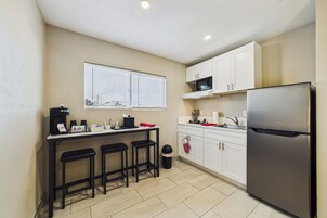 Cocina compacta con refrigerador, microondas, lavamanos y ventana con persianas. Hay dos taburetes de bar y un cubo de basura.