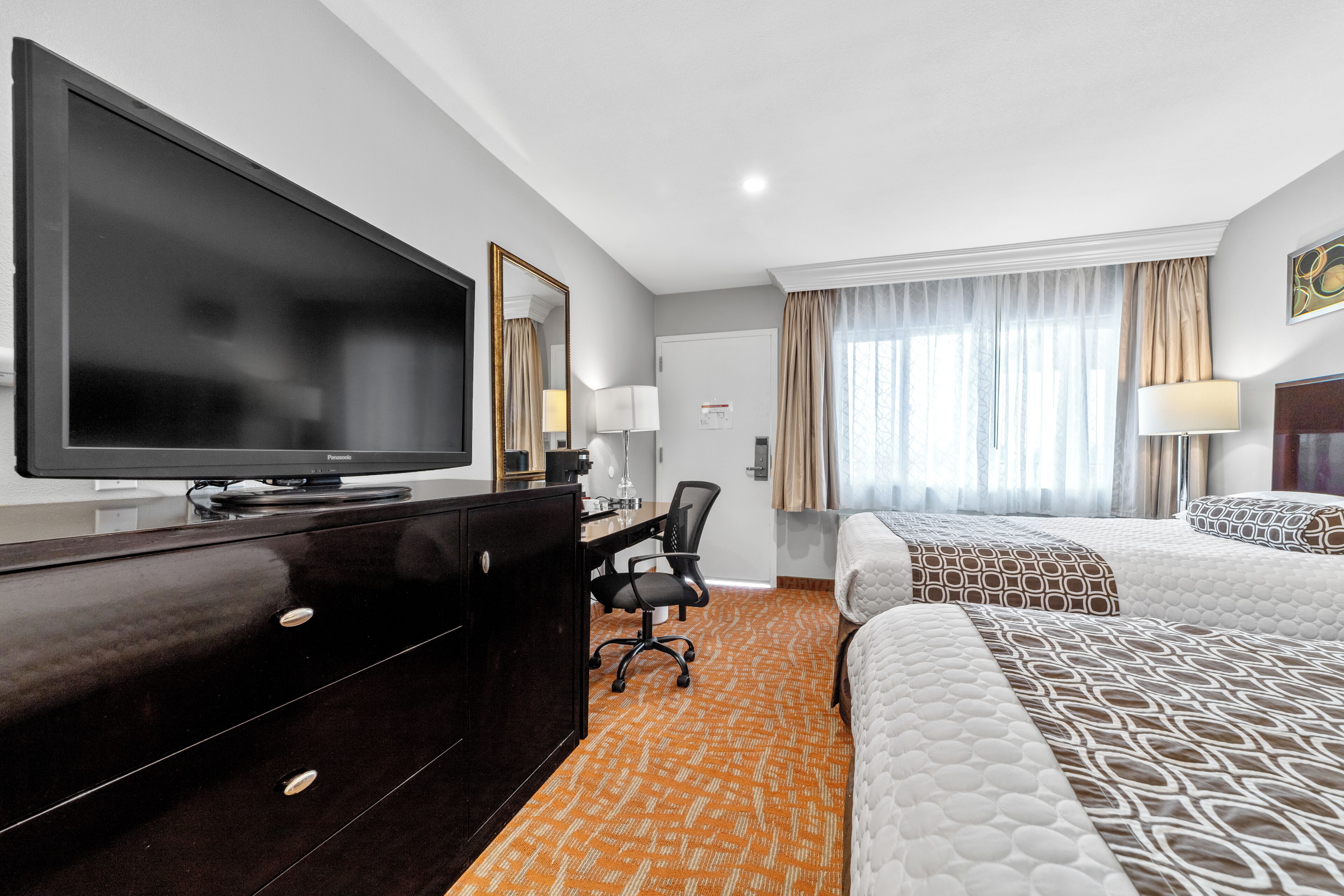 A hotel room with a large flat-screen TV, a desk with a chair, two beds with patterned bedding, and a window with curtains.