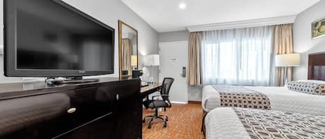 A hotel room with a large flat-screen TV, a desk with a chair, two beds with patterned bedding, and a window with curtains.