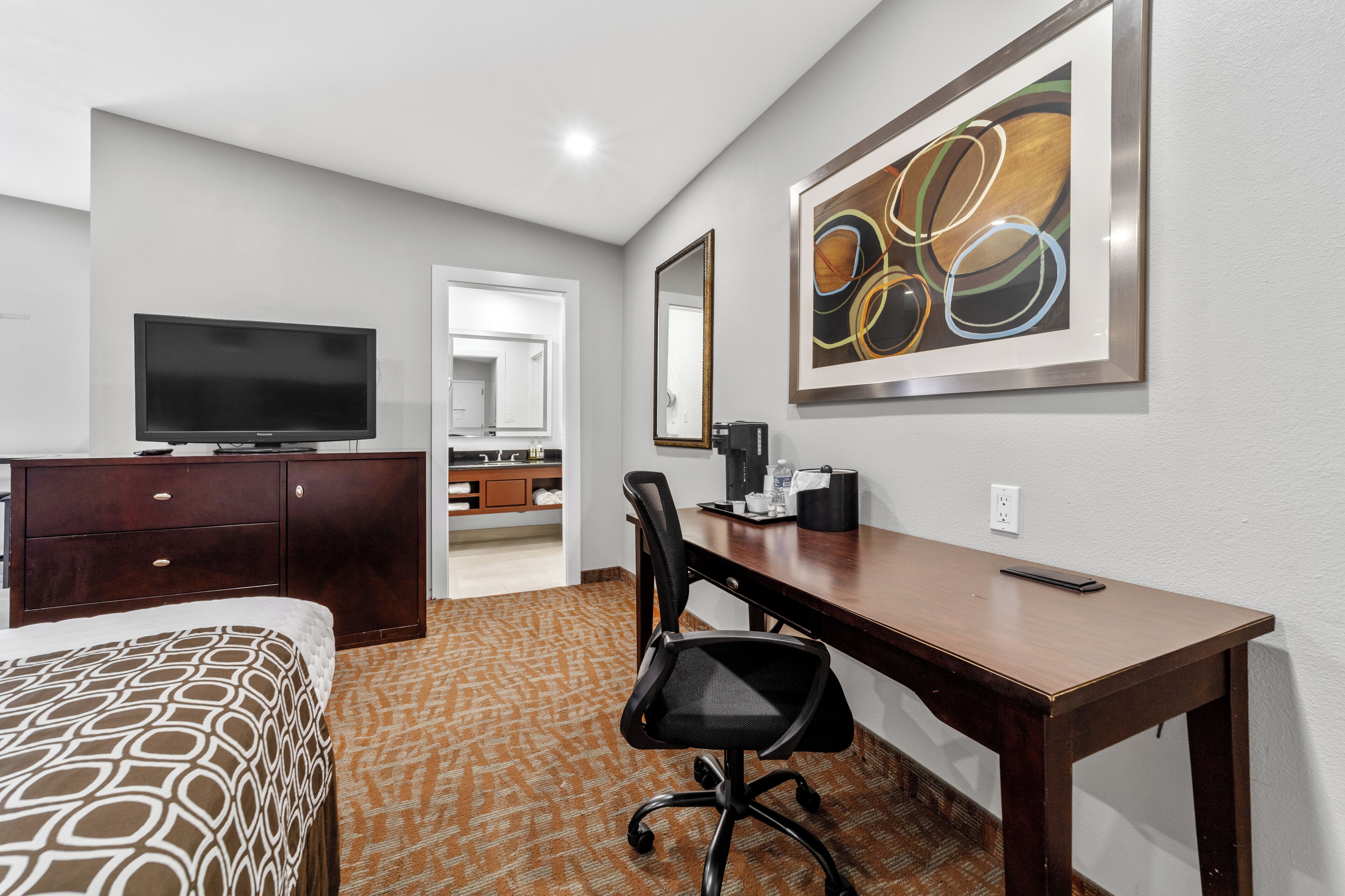 A hotel room with a bed, a desk, a chair, a television, a coffee maker, and a framed abstract painting on the wall.