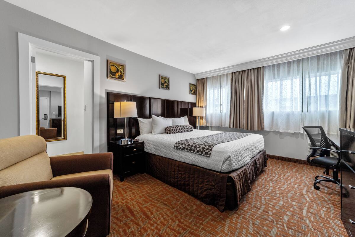 A hotel room with a bed, two bedside tables with lamps, a chair, a desk, and a large window with curtains.