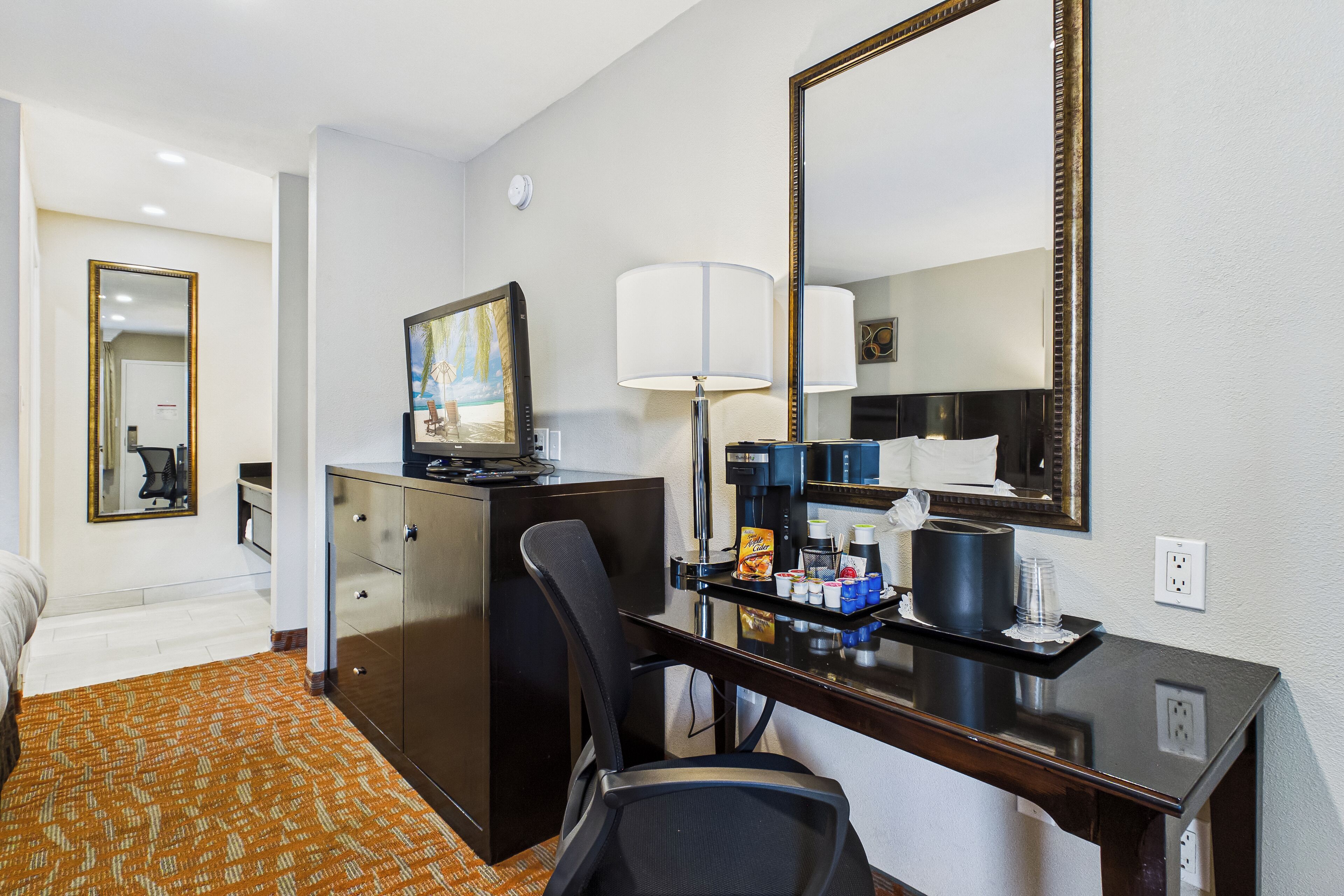 A hotel room with a bed, a desk with a lamp, a coffee maker, a painting, and a mirror.
