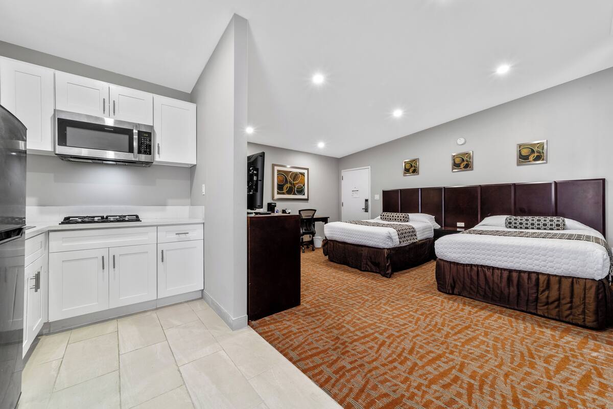A modern hotel room with a kitchenette, two beds, a large window, and a carpeted floor.