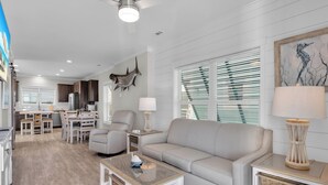 Una sala de estar moderna con un sofá, una mesa de centro y una lámpara. Hay un comedor con sillas y una mesa. El ambiente tiene piso de madera y paredes blancas.