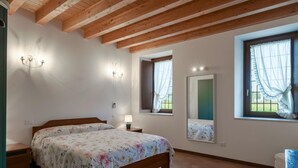 Une chambre avec un plafond en bois, un lit recouvert d’une literie à motifs floraux, deux lampes fixées au mur, une fenêtre ornée de rideaux légers et un miroir qui reflète la fenêtre.