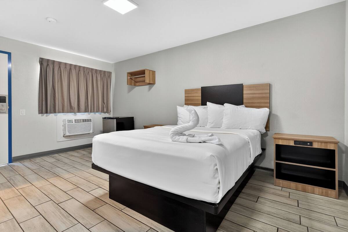 A modern hotel room with a large bed, a bedside table, a wall-mounted shelf, and a window with curtains.
