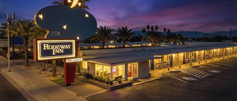 Un motel con un letrero luminoso grande, un estacionamiento y una hilera de habitaciones con ventanas visibles.