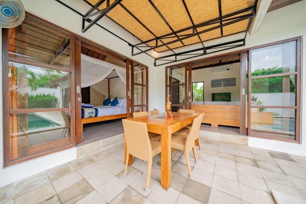 Una habitación moderna con un gran ventanal corredero que da a un balcón, una mesa de comedor de madera con sillas y una cama con dosel.