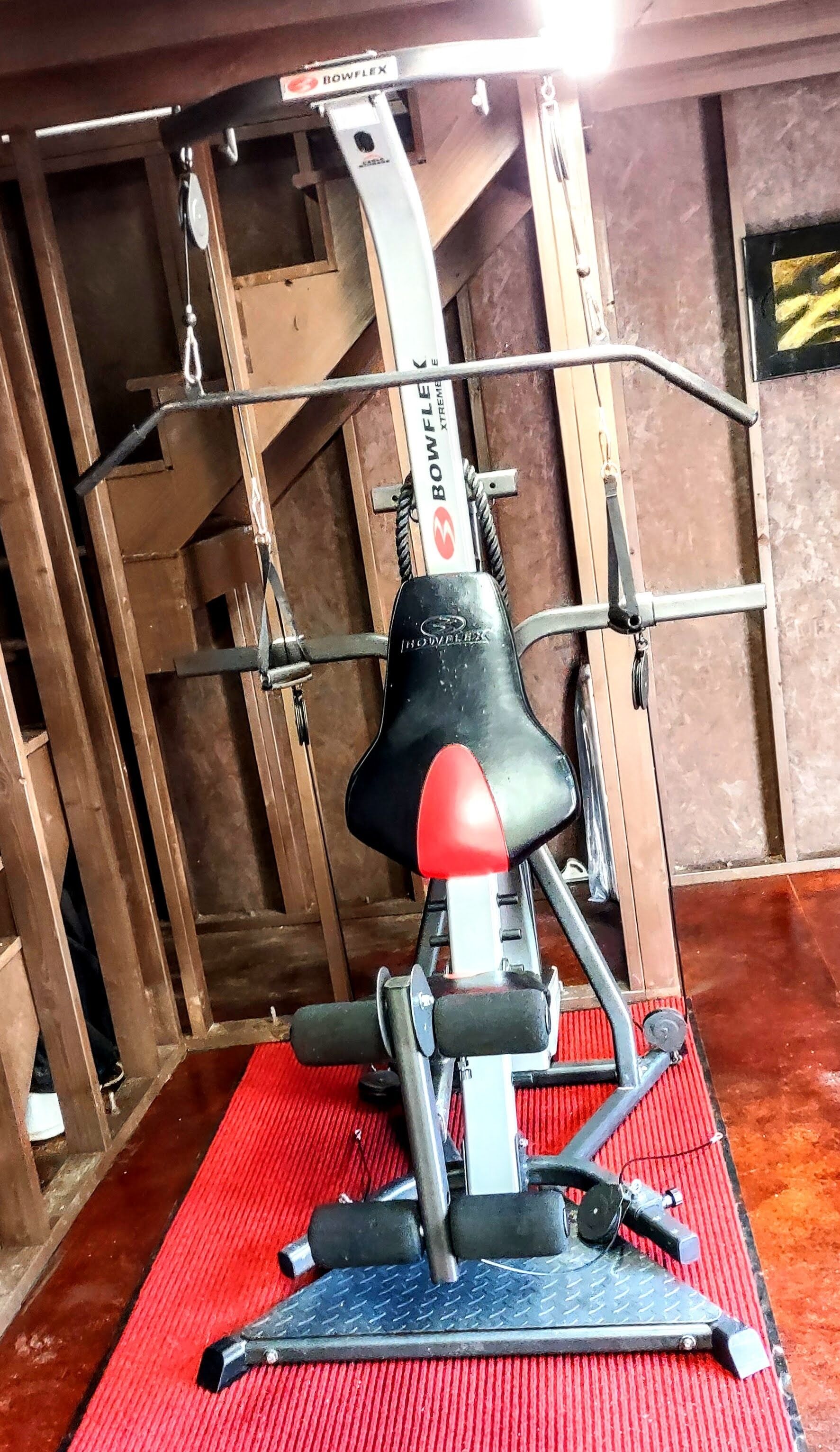 A Bowflex XtremE home gym setup with a red mat, metal frame, and black seat.