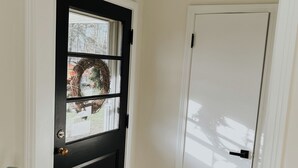 Une porte noire avec une fenêtre, une porte blanche à côté, et un paillasson marron sur un plancher en bois.