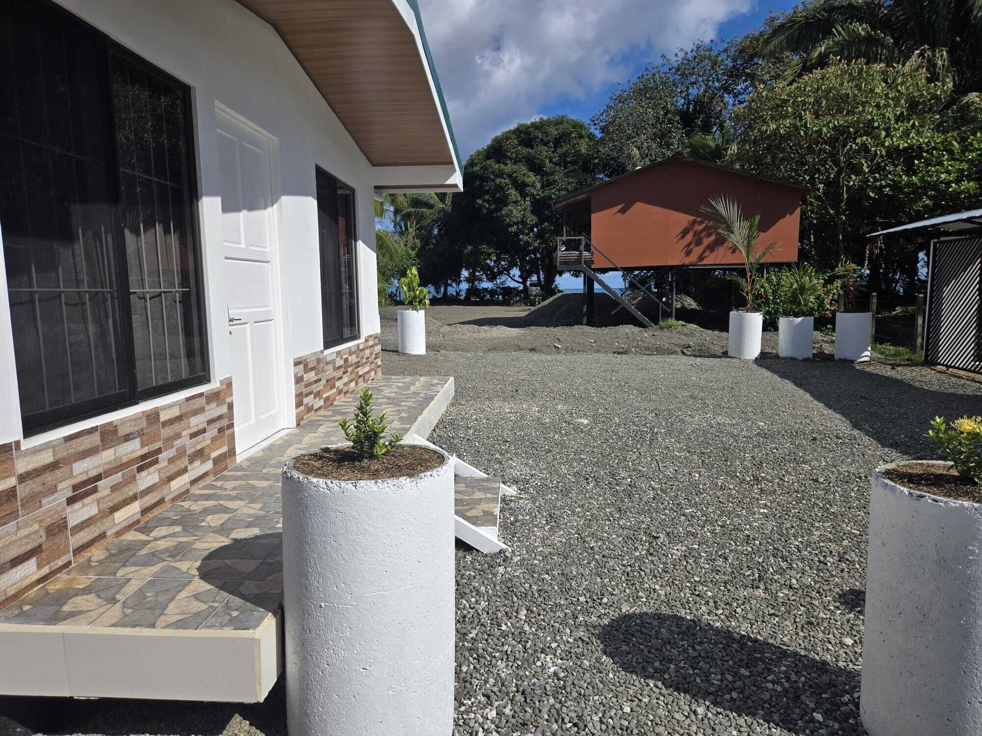 A building with a white exterior, a tiled entrance, and a gravel driveway. There are potted plants and a small structure in the background.