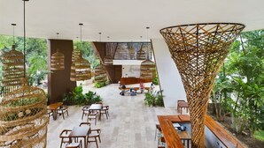 An indoor dining area with wicker chairs and tables, a central reception desk, and a large woven structure.