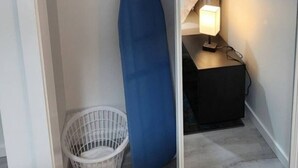 A bedroom with a bed, a bedside table with a lamp, a hanging laundry rack, a blue ironing board, and a white laundry basket.