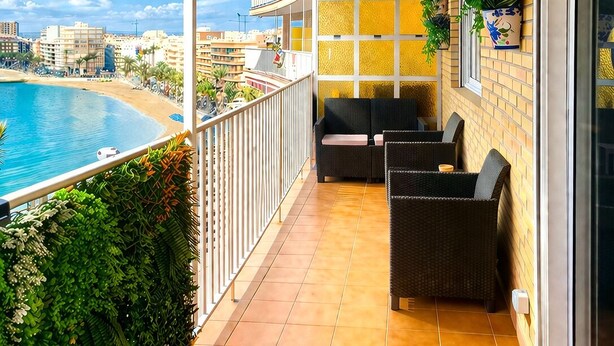 A balcony with a red awning, outdoor seating, potted plants, and a view of the beach and cityscape. - Beachfront Escape with Spacious Terrace and Stunning Seaview (Torrevieja)