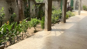 Een overdekt buitengebied met betegelde vloer, betonnen pilaren en diverse planten.