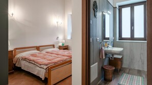 A bedroom with a wooden bed, bedside lamps, and a nightstand. Adjacent is a bathroom with a sink, mirror, and window.