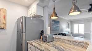 A modern kitchen with a marble countertop, stainless steel refrigerator, and two pendant lights. - Oceanfront Condo, Heated Pool, Fitness Center (Virginia Beach)