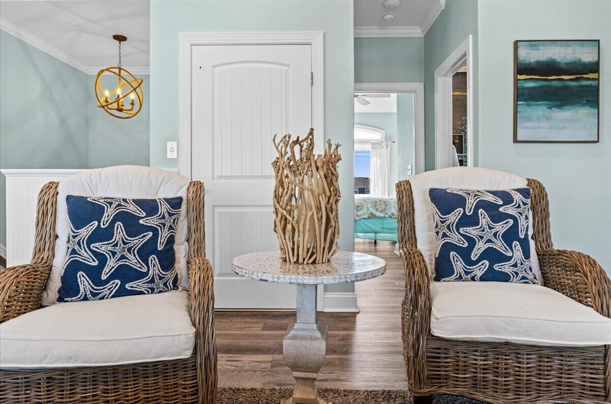 A room with two wicker chairs with star-patterned cushions, a round table with a decorative centerpiece, and a painting on the wall.