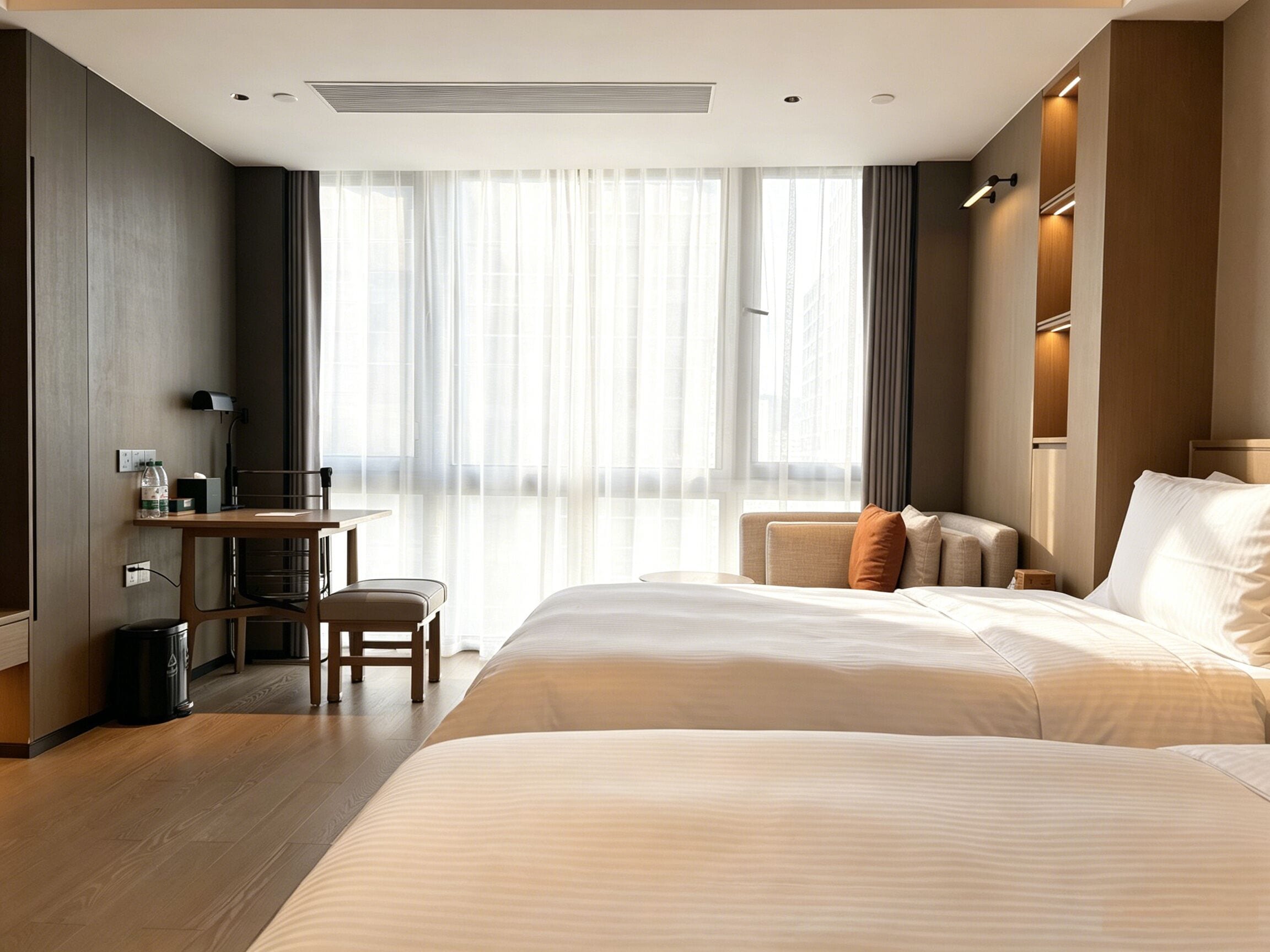 A hotel room with a large bed, a desk, a chair, and a view of the city through sheer curtains.
