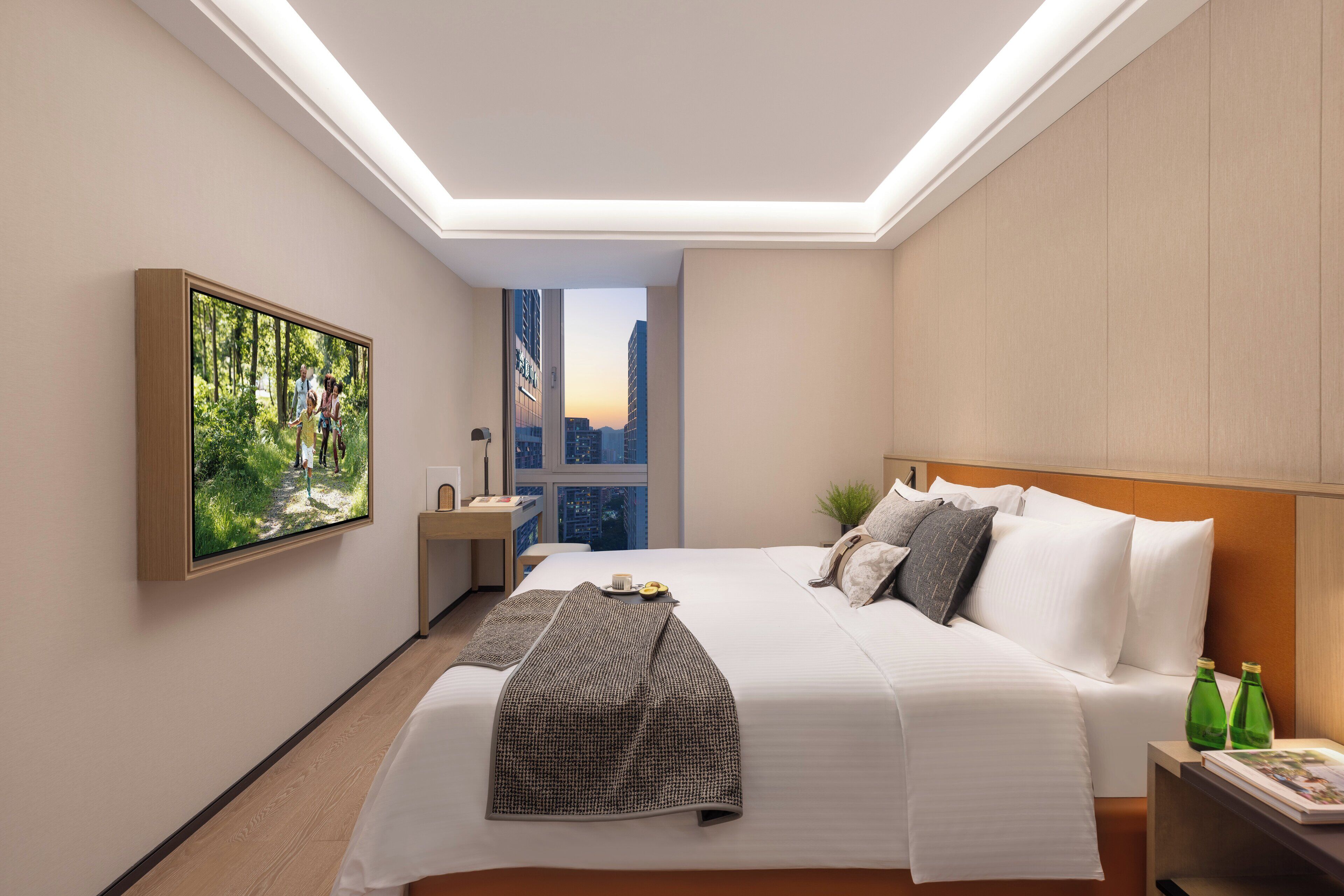 A modern hotel room with a large bed, a nightstand, a window with a city view, and a painting on the wall.