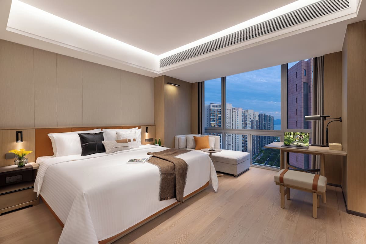 A modern hotel room with a large bed, a desk, and a view of the cityscape.