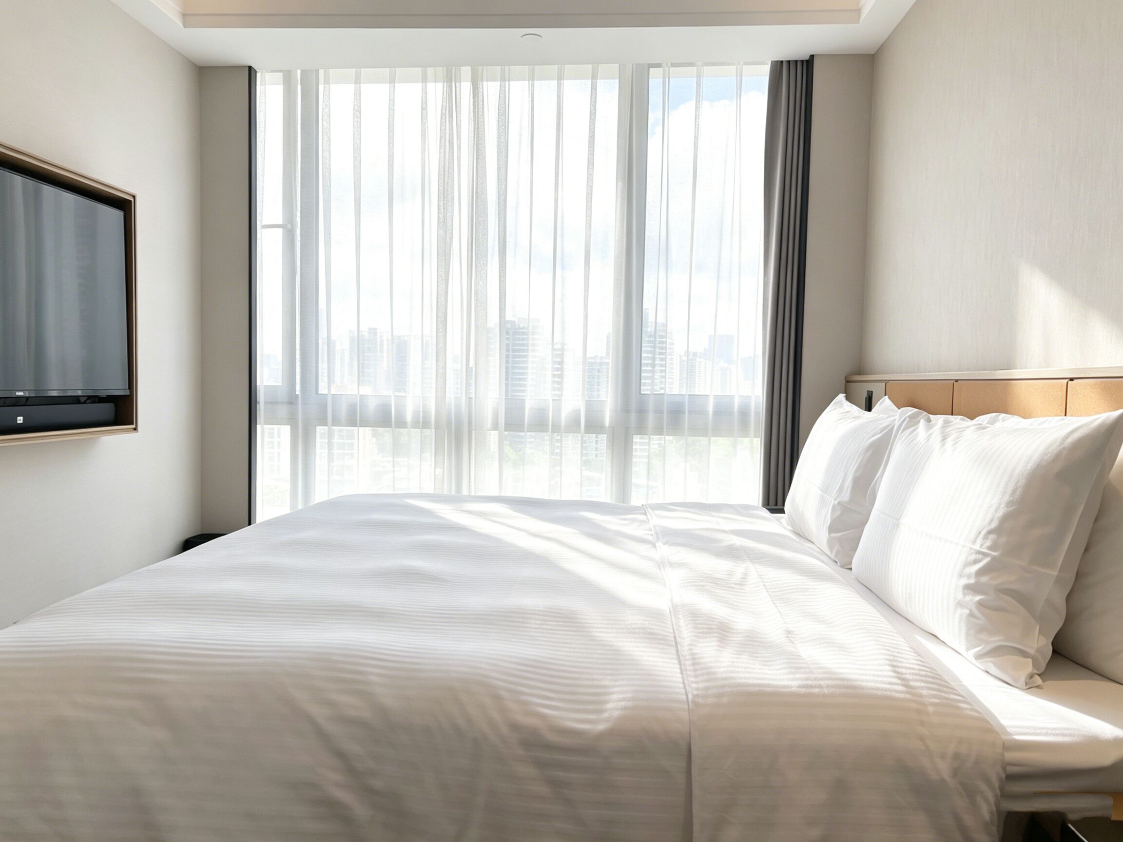 A hotel room with a bed, a TV mounted on the wall, and a view of the city through large windows.