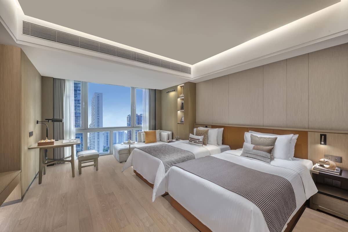 A modern hotel room with two beds, a desk, and a large window offering a city view.